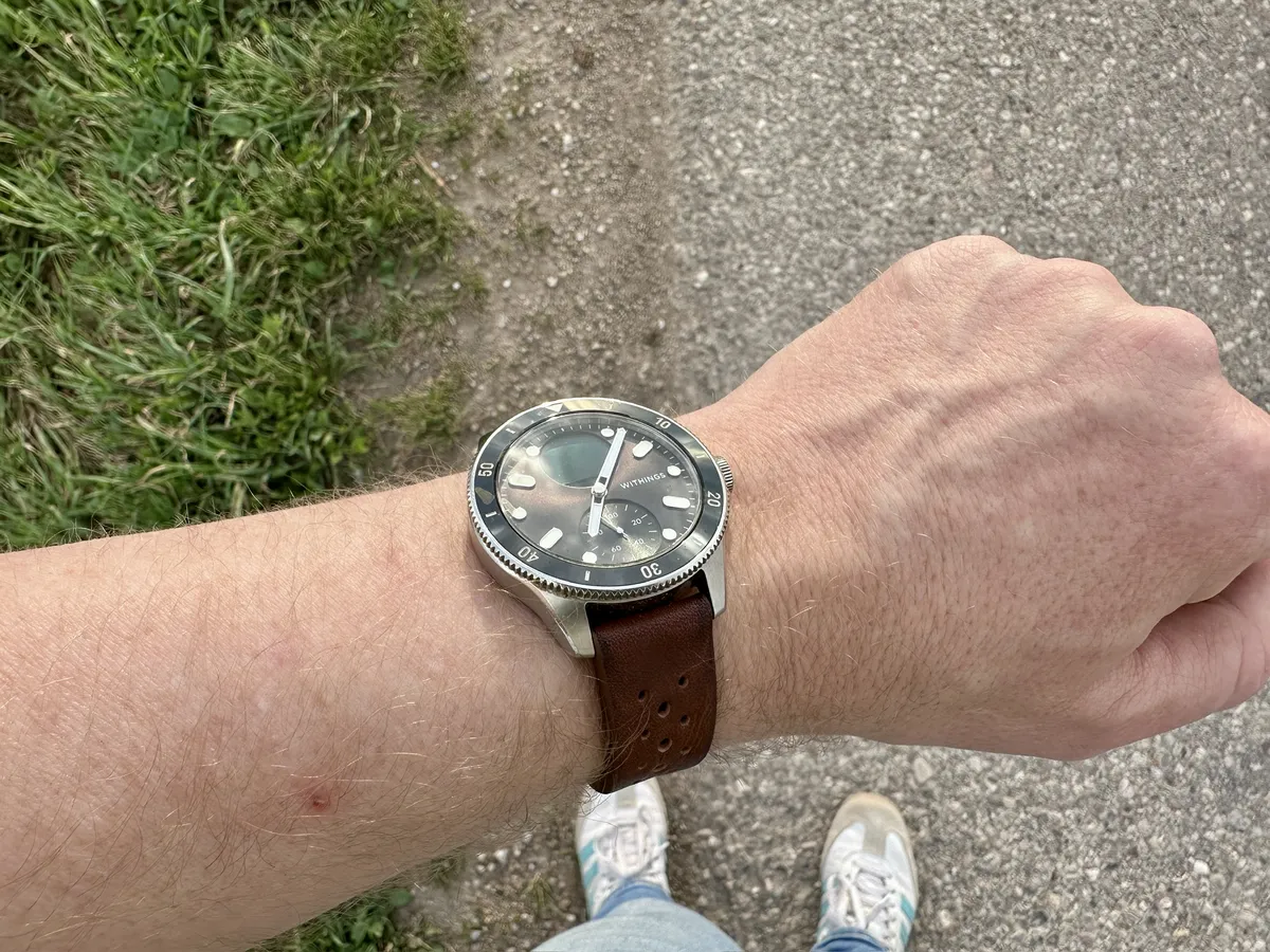Eine Hand mit einem Uhrenarmband aus braunem Leder, die eine schwarze Withings Nova mit weißen Ziffern und Zeigern trägt. Im Hintergrund ist ein Gehweg und Gras sichtbar.