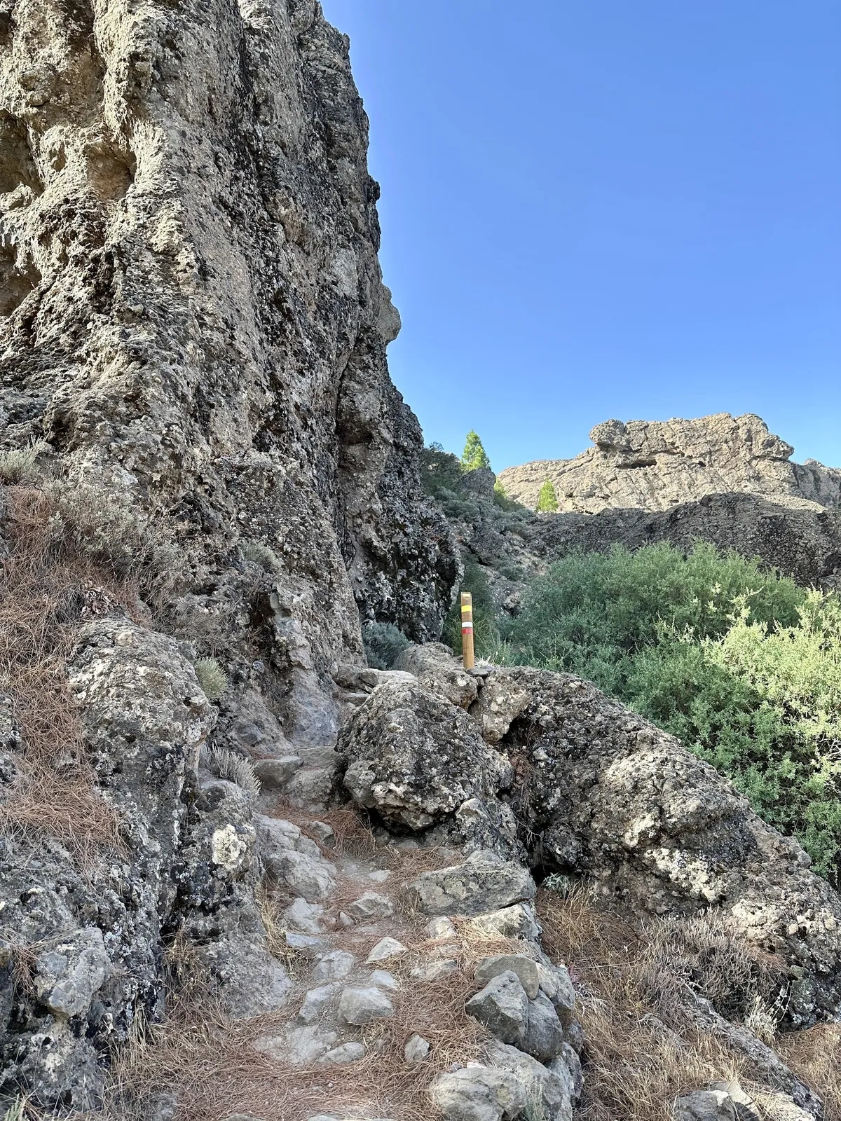 Ein schmaler Wanderweg führt zwischen felsigen Klippen und grünem Gebüsch unter einem klaren blauen Himmel hindurch.