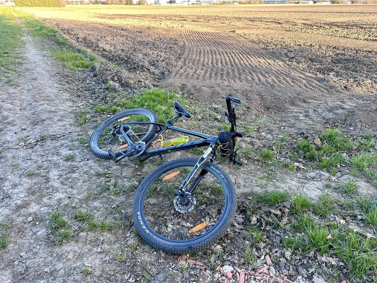 Ein schwarzes Mountainbike liegt auf einem unebenen, frisch bearbeiteten Feldweg. Im Hintergrund sind bearbeitete Felder und Wiese sichtbar.