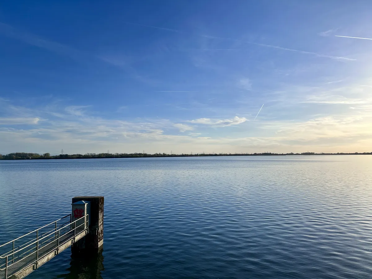 Ein ruhiger See unter einem klaren, blauen Himmel, mit sanften Wellen und einem kleinen Steg, der ins Wasser führt. Am Horizont sind Bäume und Strukturen zu erkennen.