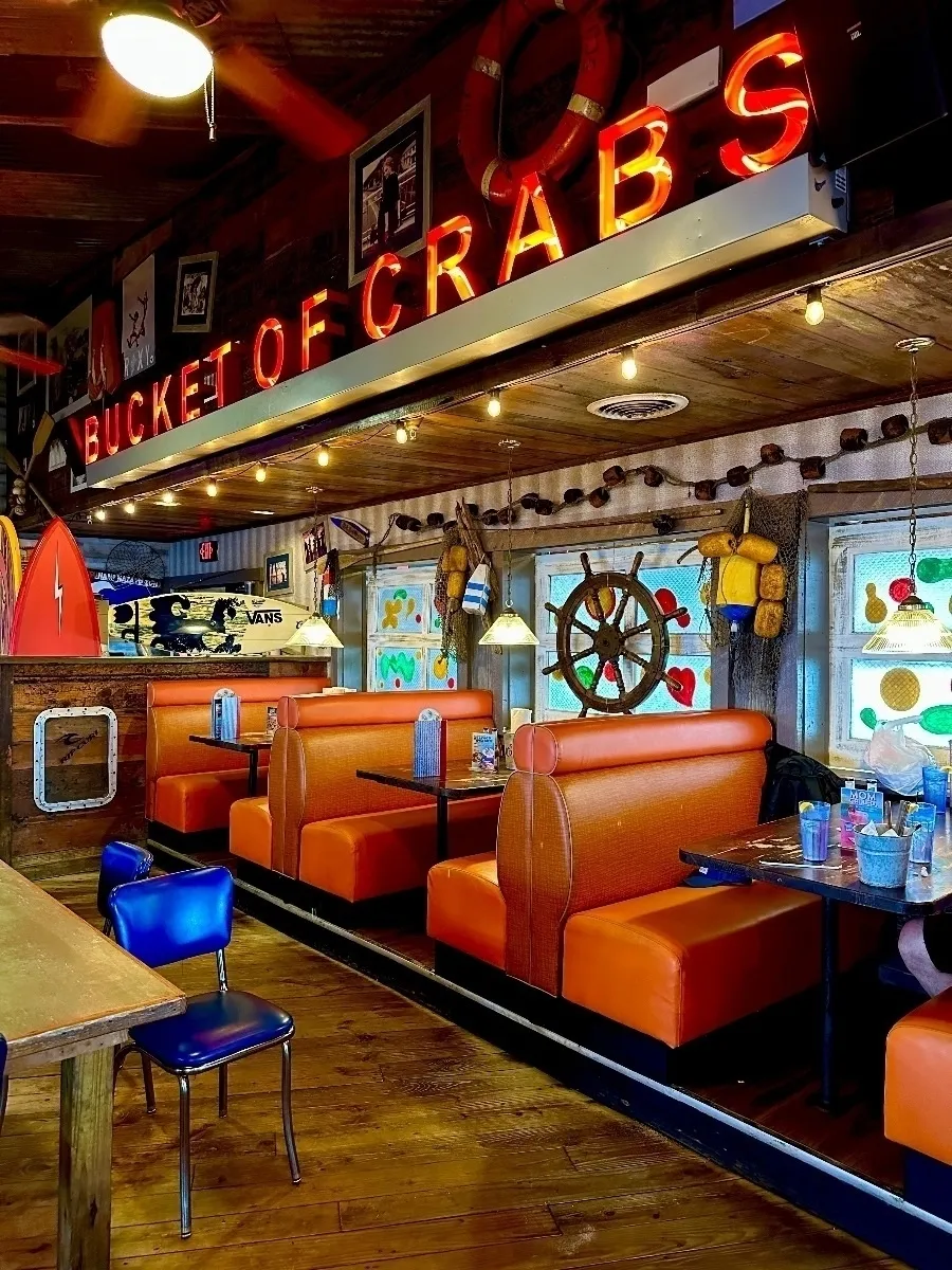 Eine Innenansicht eines rustikalen, maritim gestalteten Restaurants mit der Leuchtreklame "BUCKET OF CRABS". Es gibt orangefarbene Kunstlederbänke und Holztische, an einem Tisch steht ein blauer Stuhl.