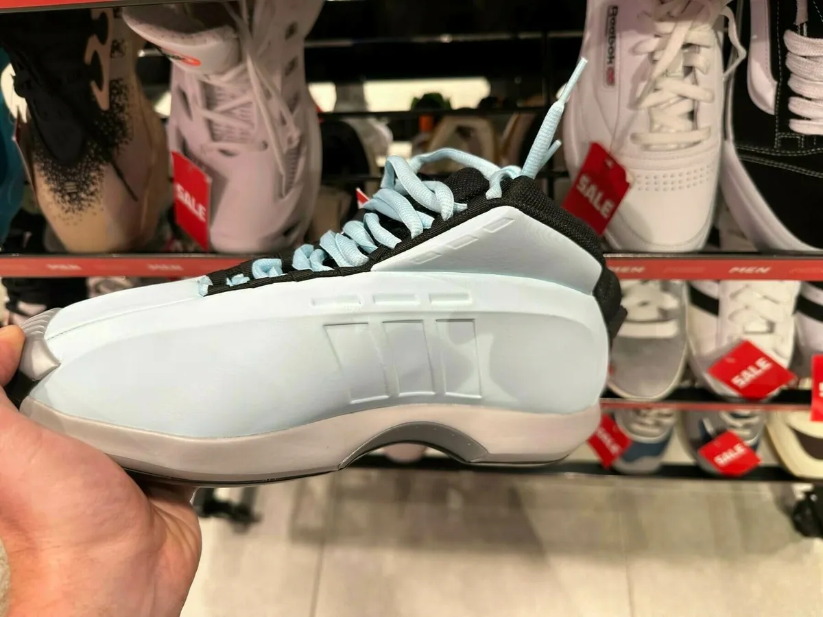 Ein hellblau-grauer Sneaker in der Hand einer Person, möglicherweise ein Modell von Adidas, mit auffälligen, hellblauen Schnürsenkeln. Im Hintergrund sind andere Sportschuhe in einem Verkaufsregal zu sehen.