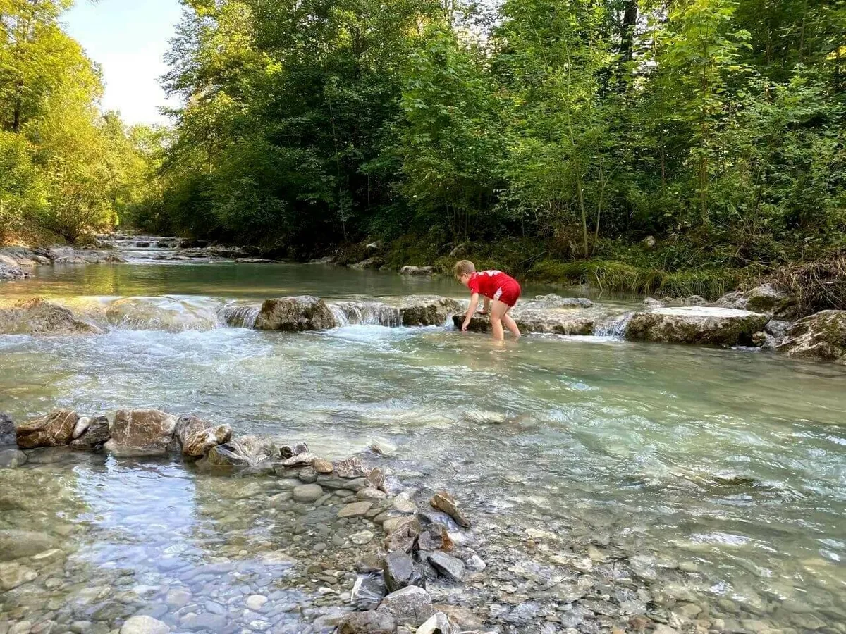 Ein Junge in roter Kleidung steht barfuß in einem klaren Bergbach mit felsigen Stufen und spielt mit Steinen im seichten Wasser.