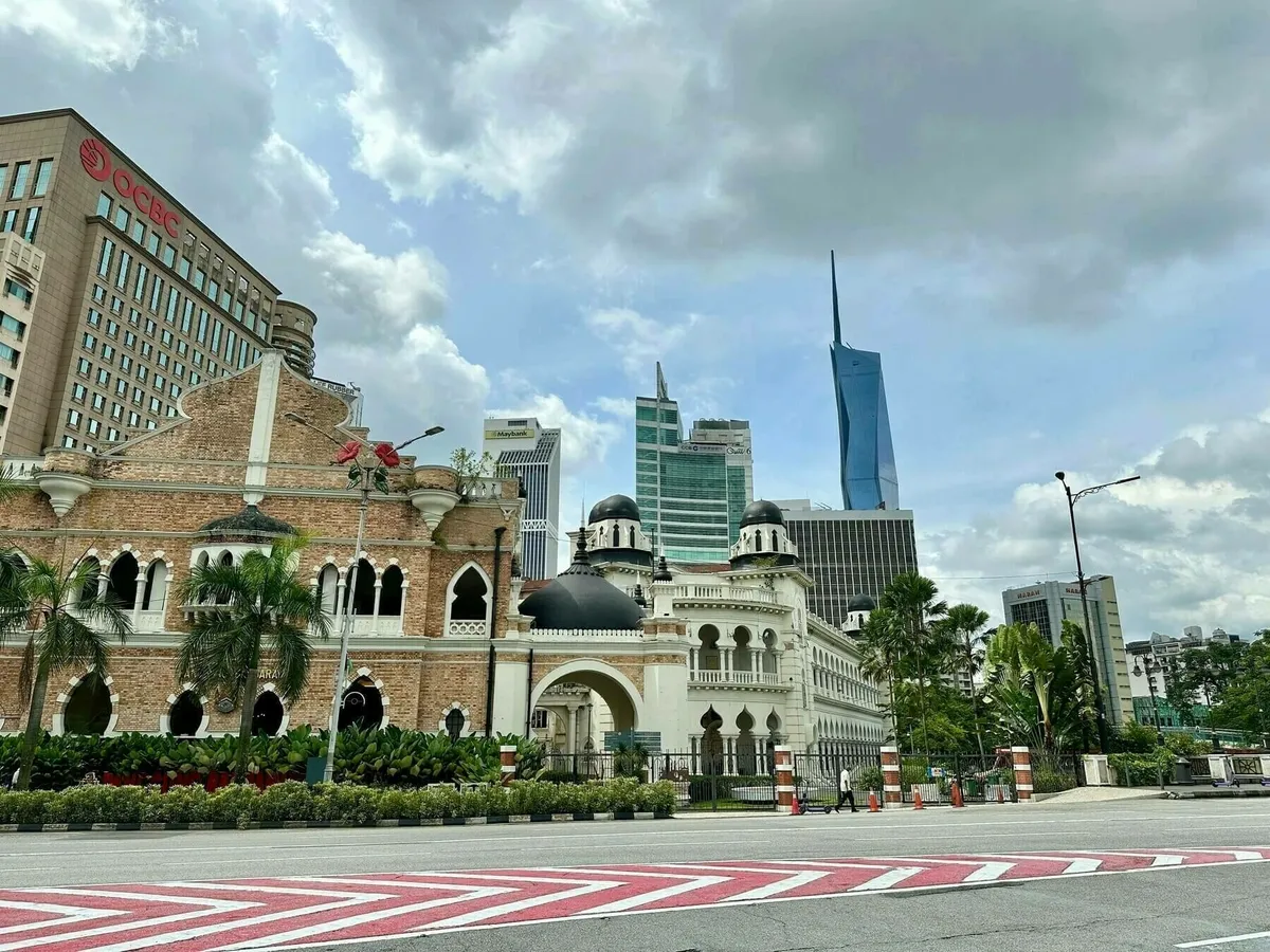 Ein Straßenblick in Kuala Lumpur, der historische Architektur und moderne Wolkenkratzer gegenüberstellt. Im Vordergrund steht ein Gebäude im maurischen Stil mit Kuppeln und Ziegeln, dahinter moderne Hochhäuser, darunter der blaue, spitze The Exchange 106 Turm.