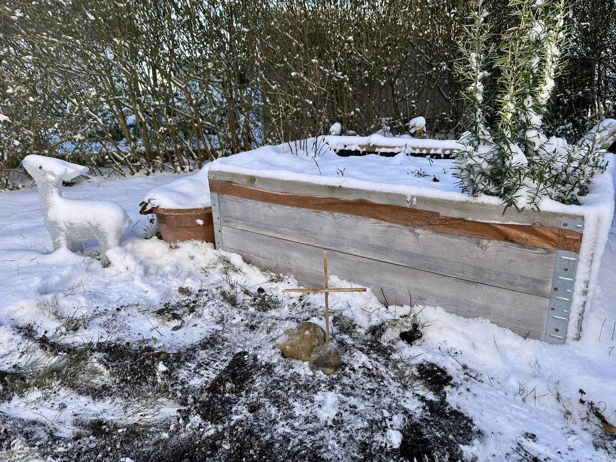 Ein schneebedeckter Gartenbereich mit einem Holzbeet und einem Kreuz aus einem Holzstab sowie einem Stein in der Erde. Im Hintergrund ist eine weiß gefrorene Landschaft mit Sträuchern zu sehen.