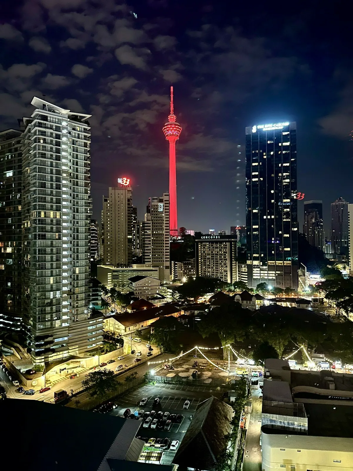 Eine nächtliche Aufnahme der Skyline von Kuala Lumpur von oben. Die beleuchtete KL Tower sticht in leuchtendem Rot heraus. Im Vordergrund sind moderne Hochhäuser und darunter ein beleuchteter Stadtpark und ein Parkplatz zu sehen.
