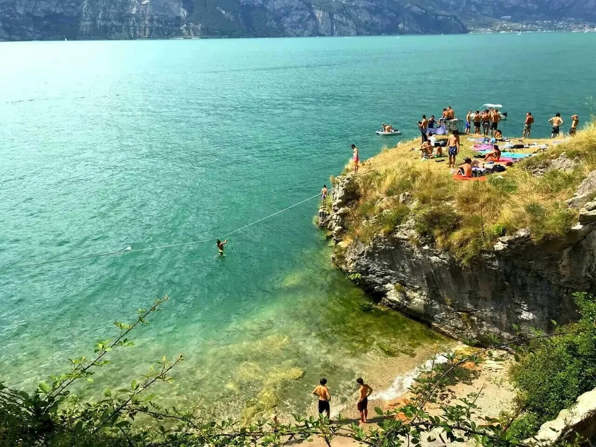 Eine felsige Bucht mit türkisblauem Wasser, vermutlich am Gardasee. Oben auf dem Felsen sonnen sich Jugendliche, und eine Person balanciert über eine gespannte Leine über dem Wasser (Slackline).