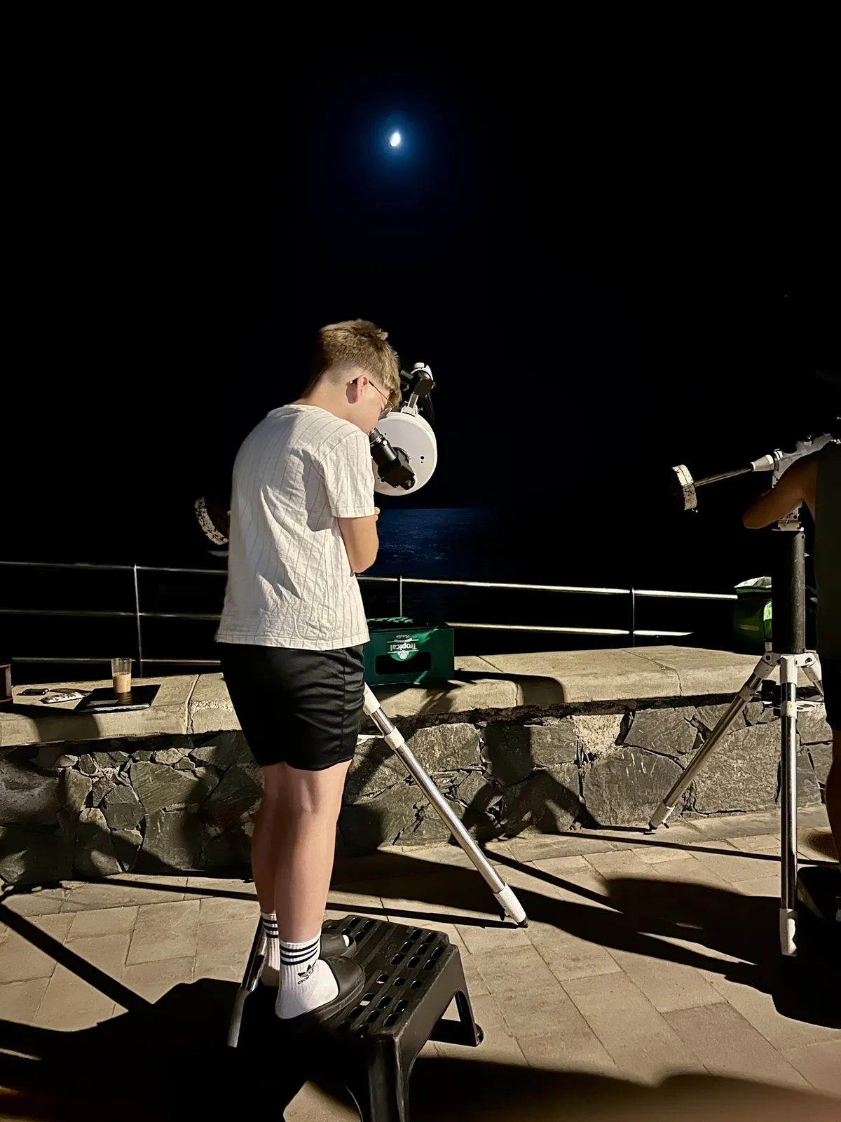 Ein Junge beobachtet nachts den Himmel durch ein Teleskop, während der Halbmond über dem Meer leuchtet.