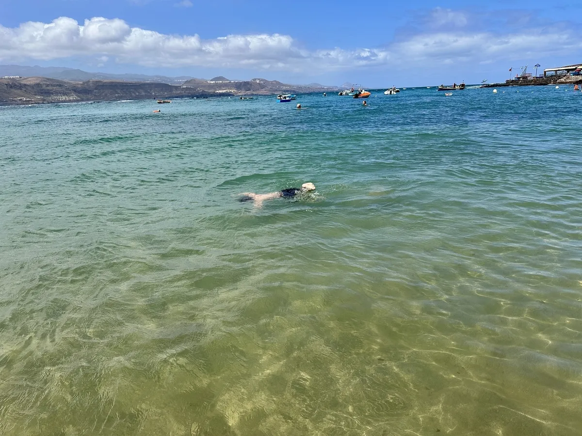 Eine Person schwimmt im klaren, türkisfarbenen Wasser eines Strandes, während Boote in der Ferne zu sehen sind.