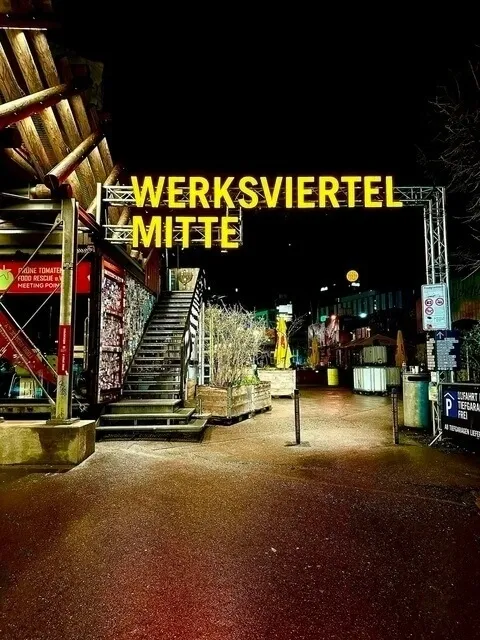 Nachtaufnahme des Eingangs zum "WERKSVIERTEL MITTE". Ein großes, gelb leuchtendes Schild überspannt eine Treppe, die zu einem geschlossenen/dunklen Bereich führt. Der Boden ist nass und reflektiert das Licht.