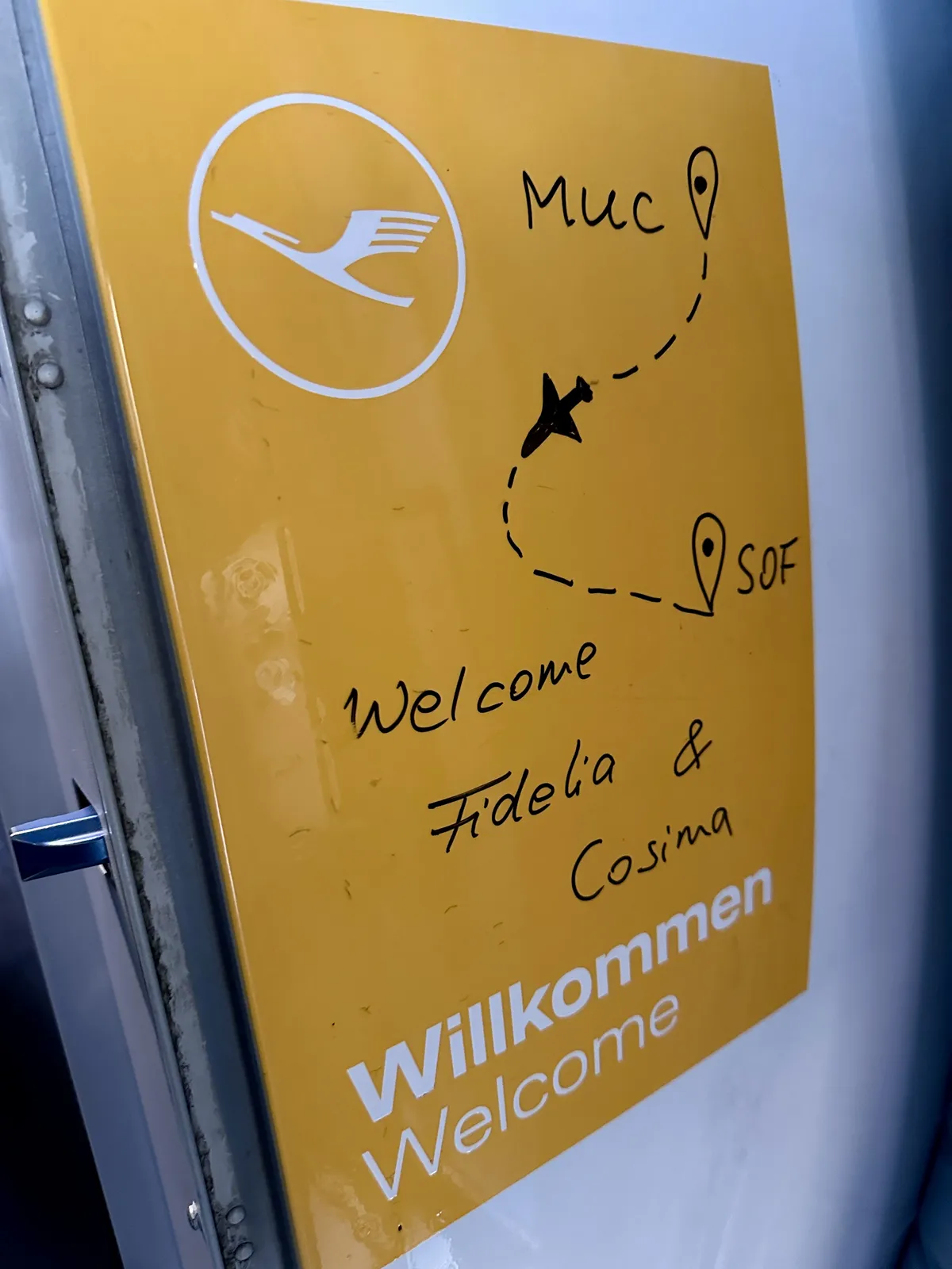 Ein gelbes Schild mit dem Logo der Lufthansa, das die Städte München (MUC) und Sofia (SOF) miteinander verbindet. Handgeschriebene Grüße an "Fidelia & Cosima" sind ebenfalls darauf zu sehen.