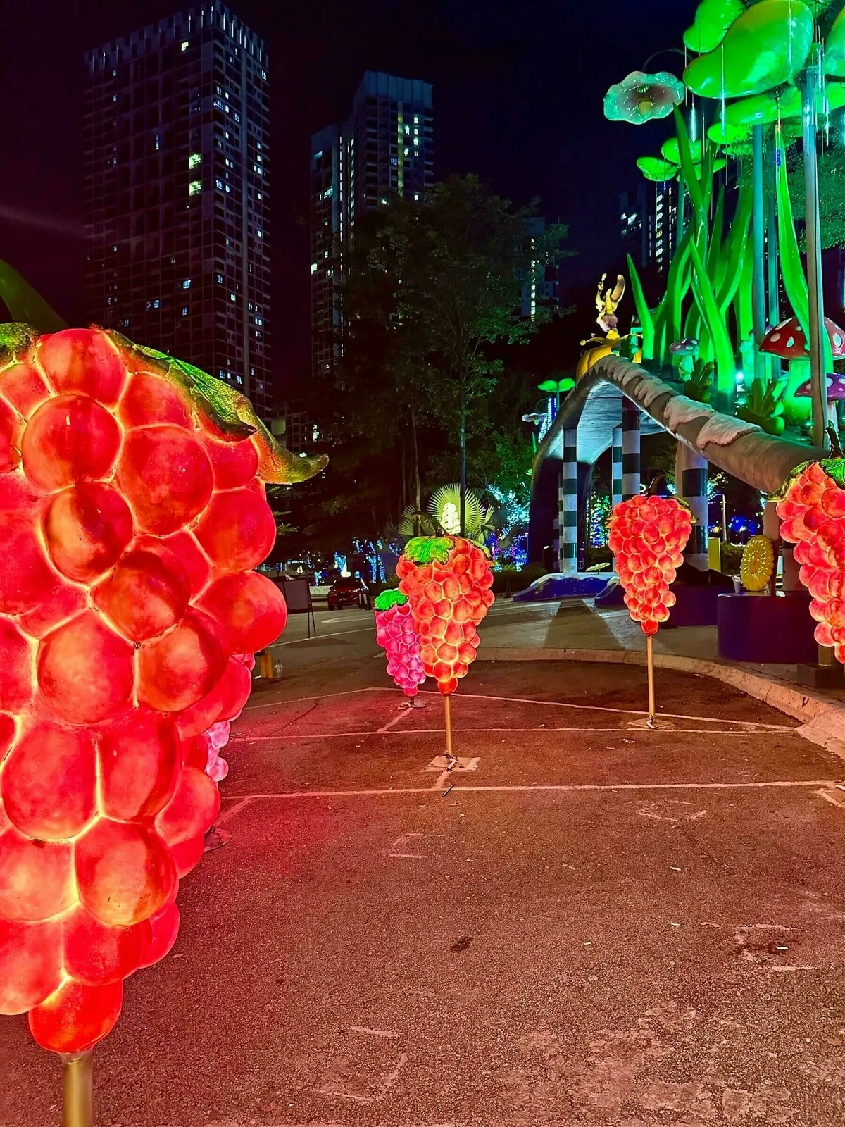 Leuchtende Traubenskulpturen säumen einen Weg nahe hoher Gebäude bei Nacht, im Hintergrund helle grüne Lichtinstallationen.