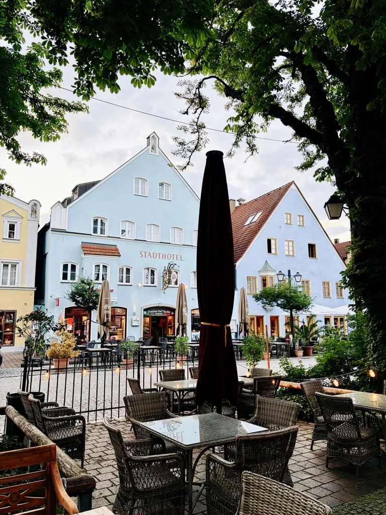 Ein malerischer Stadtplatz mit einem gelben und einem blauen Gebäude, das ein Hotel beherbergt. Auf der Terrasse stehen Tische und Stühle aus Rattan, umgeben von Bäumen und einer gemütlichen Atmosphäre.