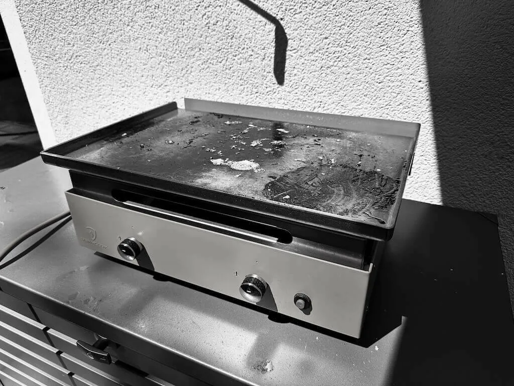 Auto-generated description: Eine Plancha mit zwei Reglern steht auf einer Metalloberfläche und zeigt Gebrauchsspuren.