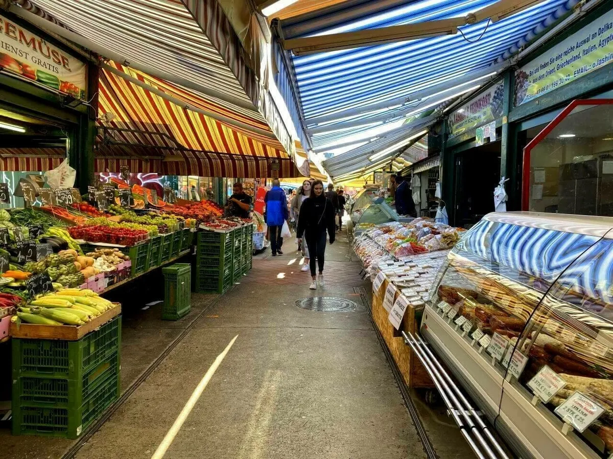 Ein belebter Marktgang auf dem Wiener Naschmarkt unter gestreiften Markisen, gesäumt von Ständen mit frischem Gemüse und einer Kühltheke mit Delikatessen.