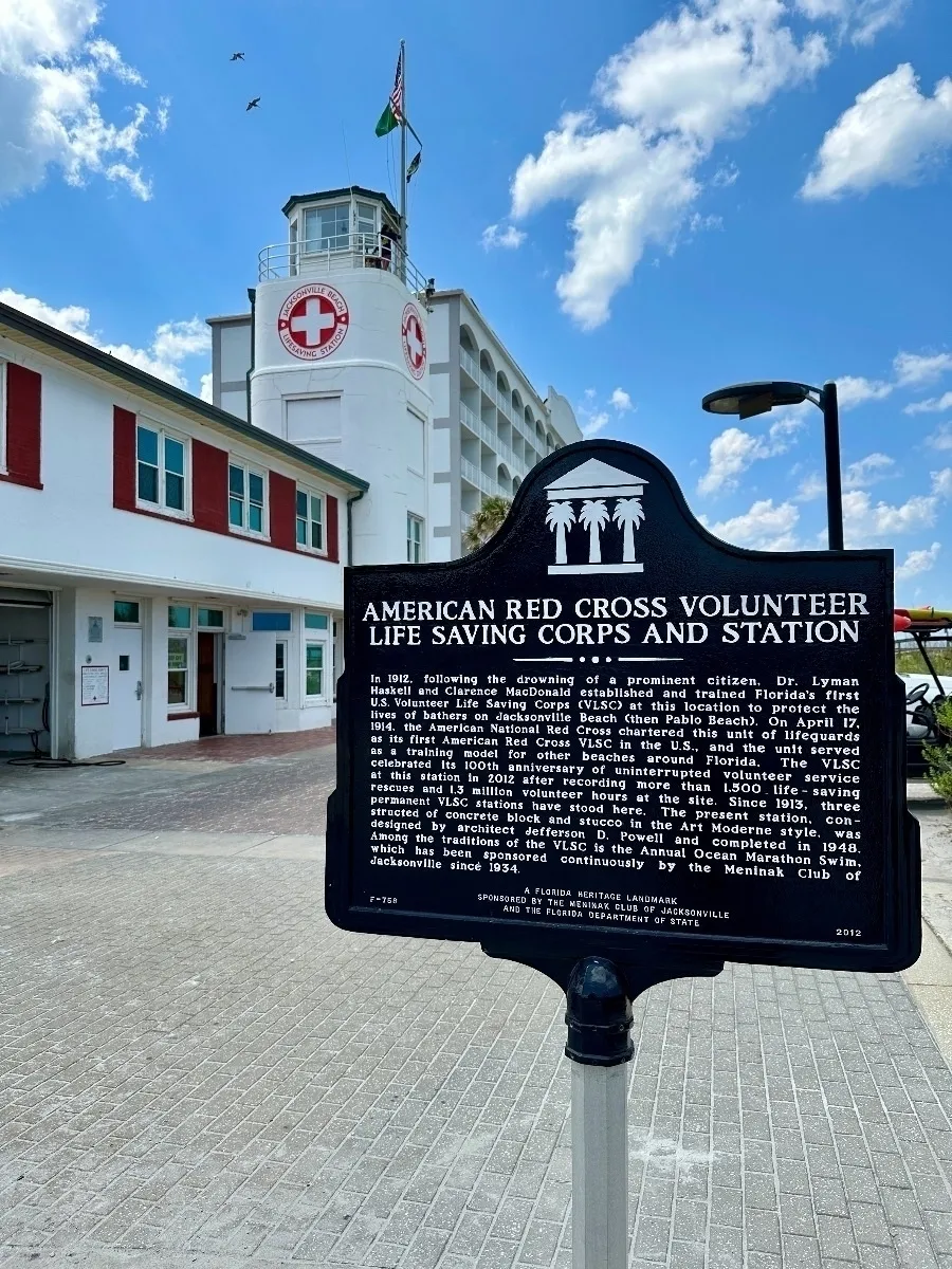Eine schwarze, historische Gedenktafel des American Red Cross Volunteer Life Saving Corps and Station vor dem weißen, turmartigen Gebäude der Rettungsstation unter einem bewölkten blauen Himmel.