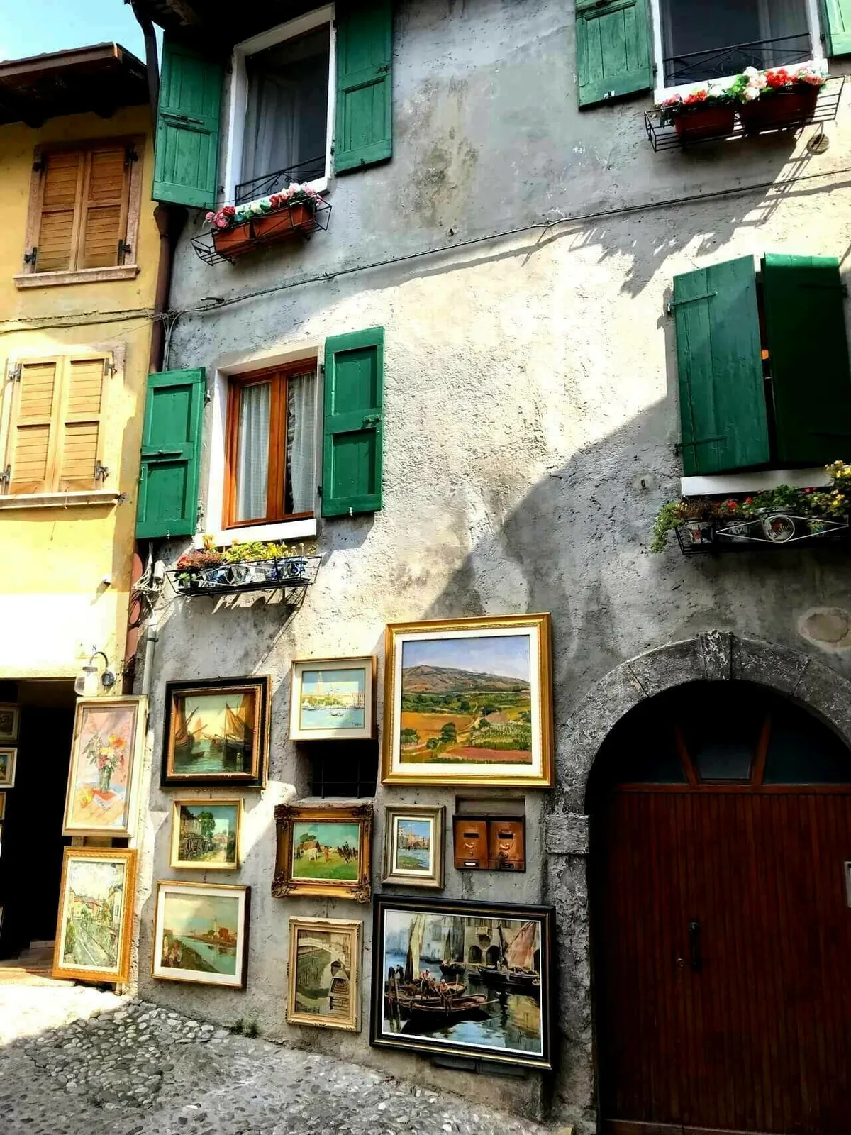 Eine rustikale Hauswand mit grünen Fensterläden, an der zahlreiche gerahmte Gemälde aufgehängt sind. Die Kunstwerke zeigen verschiedene Landschaften und Seestücke.