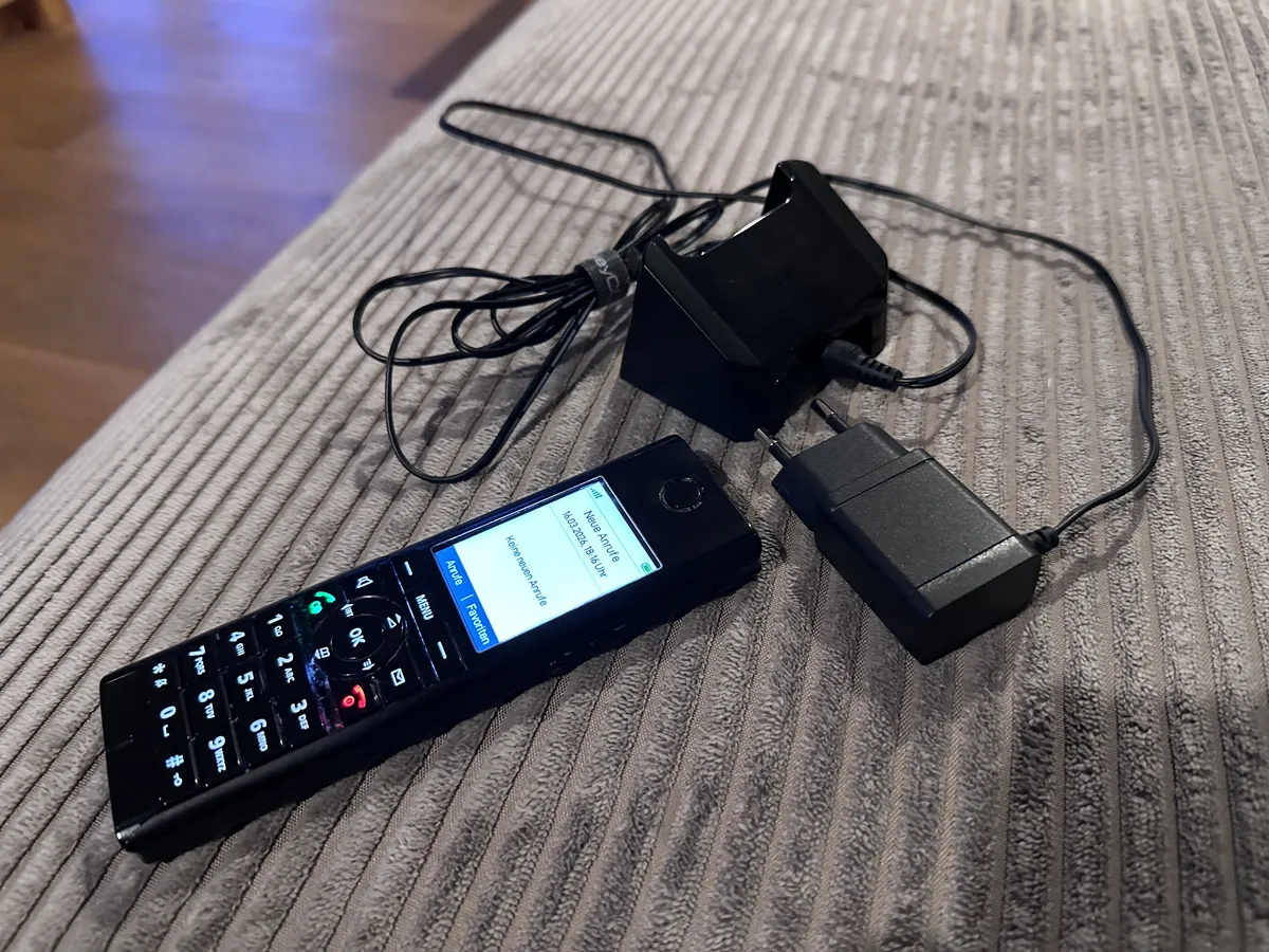 Ein mobiles Festnetztelefon liegt auf einem gestreiften Sofa, neben einem Ladekabel und einer Ladeschale. Der Bildschirm des Handys zeigt eine Nachricht an.