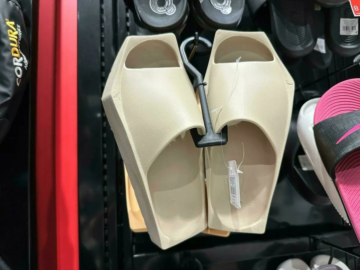 Ein Paar beigefarbene oder sandfarbene Badelatschen (Slides) mit einem kantigen, ungewöhnlichen Design, aufgehängt in einem Geschäft neben anderen Schuhen.