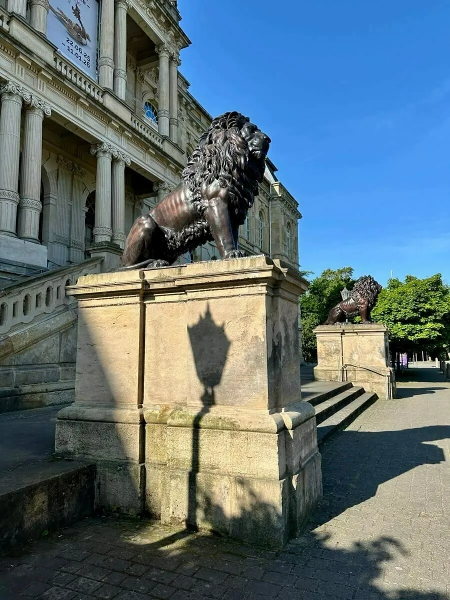 Vor dem herzoglichen Museum stehen zwei Löwenstatuen auf steinernen Podesten, während die Sonne einen klaren Schatten wirft.