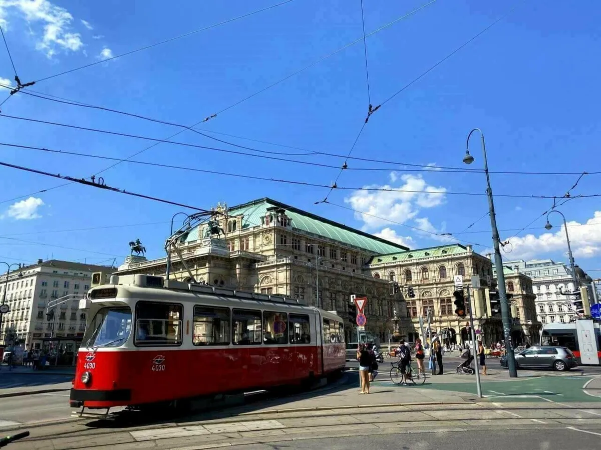 Eine rote Straßenbahn der Wiener Linien an einer Kreuzung vor dem historischen Gebäude der Wiener Staatsoper unter blauem Himmel.