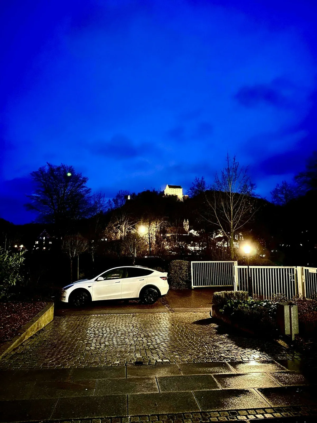 Ein weißes Tesla Model Y parkt auf einem nassen Kopfsteinpflasterweg, umgeben von Bäumen und der  beleuchteten Rosenburg  in der Ferne, unter einem tiefblauen Abendhimmel.
