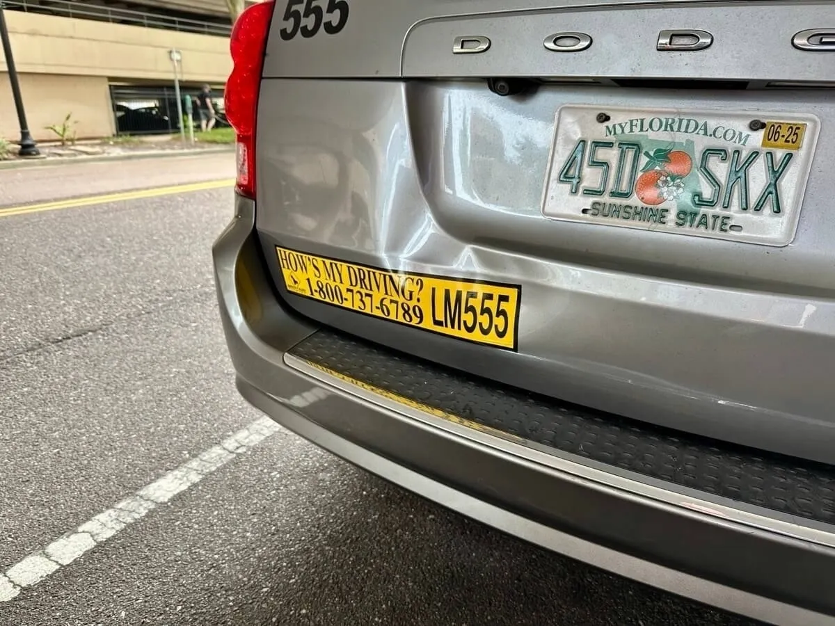 Die Heckansicht eines silbernen Minivans der Marke Dodge mit einer leichten Delle im Kofferraumdeckel. Es ist ein Nummernschild aus Florida (MyFlorida.com, SUNSHINE STATE) mit der Kennzeichnung 450 SKX angebracht, gültig bis 06/25. Direkt unter dem Kennzeichen befindet sich ein gelb-schwarzer Aufkleber mit der Aufschrift "HOW'S MY DRIVING? 1-800-737-6789" und einer eindeutigen Kennung LM555. Oben links auf der Karosserie steht die Nummer 555.