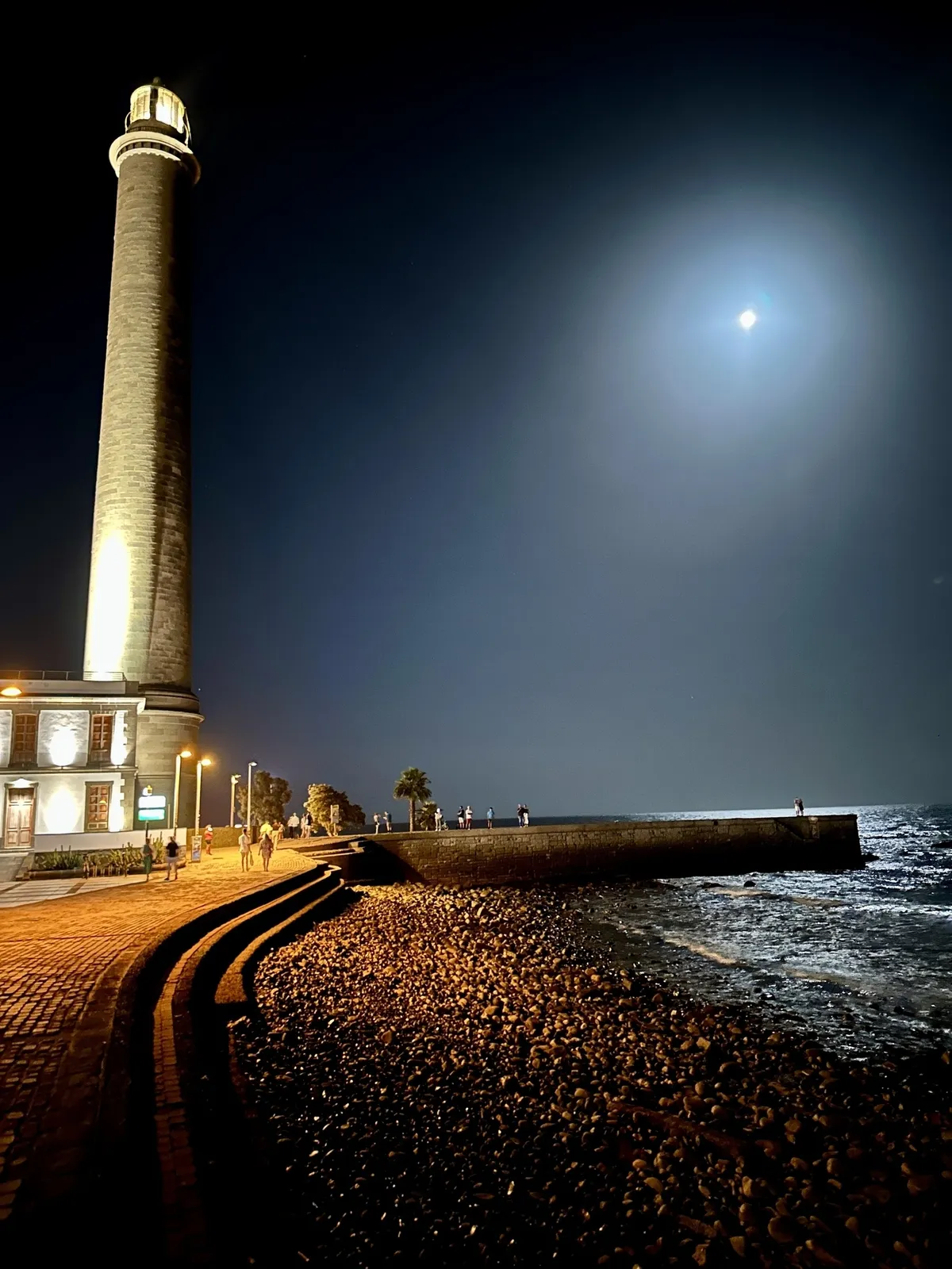 Ein beleuchteter Leuchtturm steht an der Küste von Maspalomas in Gran Canaria unter einem klaren Nachthimmel mit Mondschein.