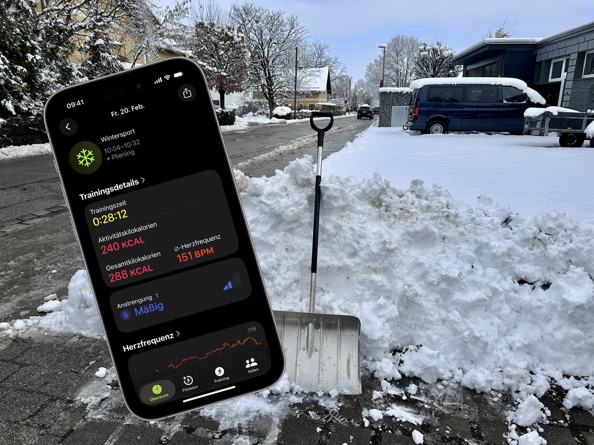 Ein Smartphone mit Trainingsdetails steht auf dem Bürgersteig neben einem Schneeschieber. Im Hintergrund ist eine verschneite Straße und ein Wohngebiet zu sehen, während Schnee auf einem kleinen Haufen angehäuft ist.