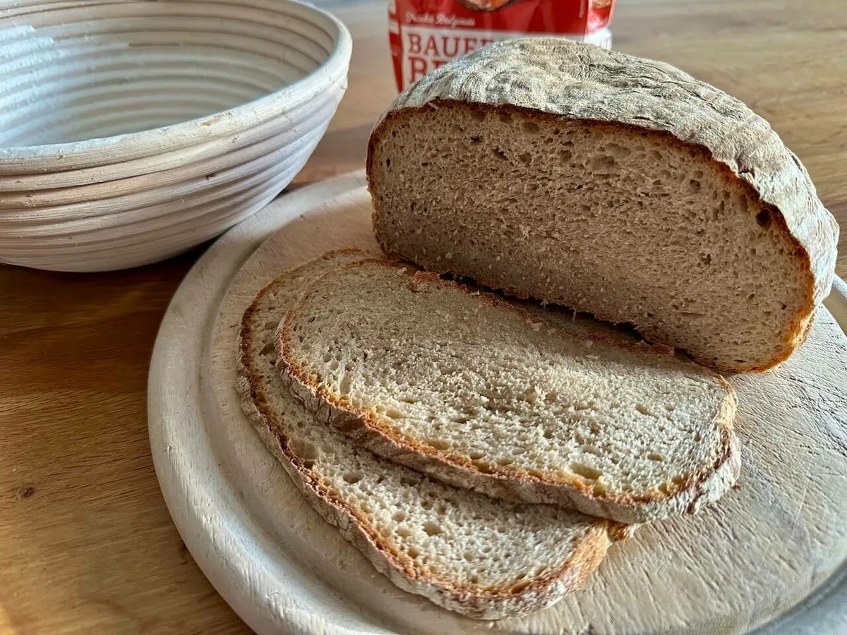 Ein aufgeschnittenes Laib Brot liegt mit mehreren Brotscheiben auf einem Holzbrett neben einer Schüssel.