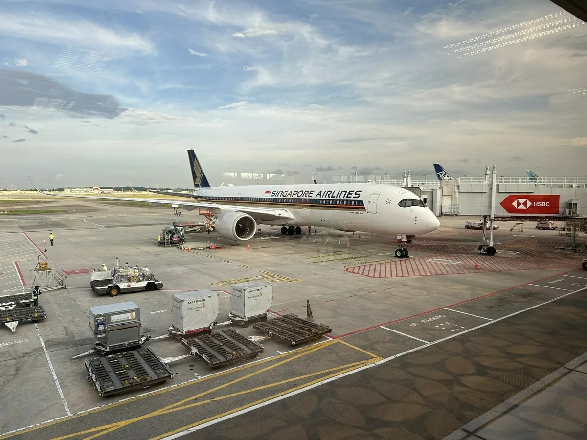 Ein Flugzeug der Singapore Airlines (vermutlich ein Airbus A350) steht auf dem Rollfeld eines Flughafens, verbunden mit einem Fluggastbrücke, die das HSBC-Logo trägt. Vor dem Flugzeug sind Gepäck- und Frachtcontainer sowie Bodenfahrzeuge zu sehen, aufgenommen durch ein großes Fenster der Abflughalle.