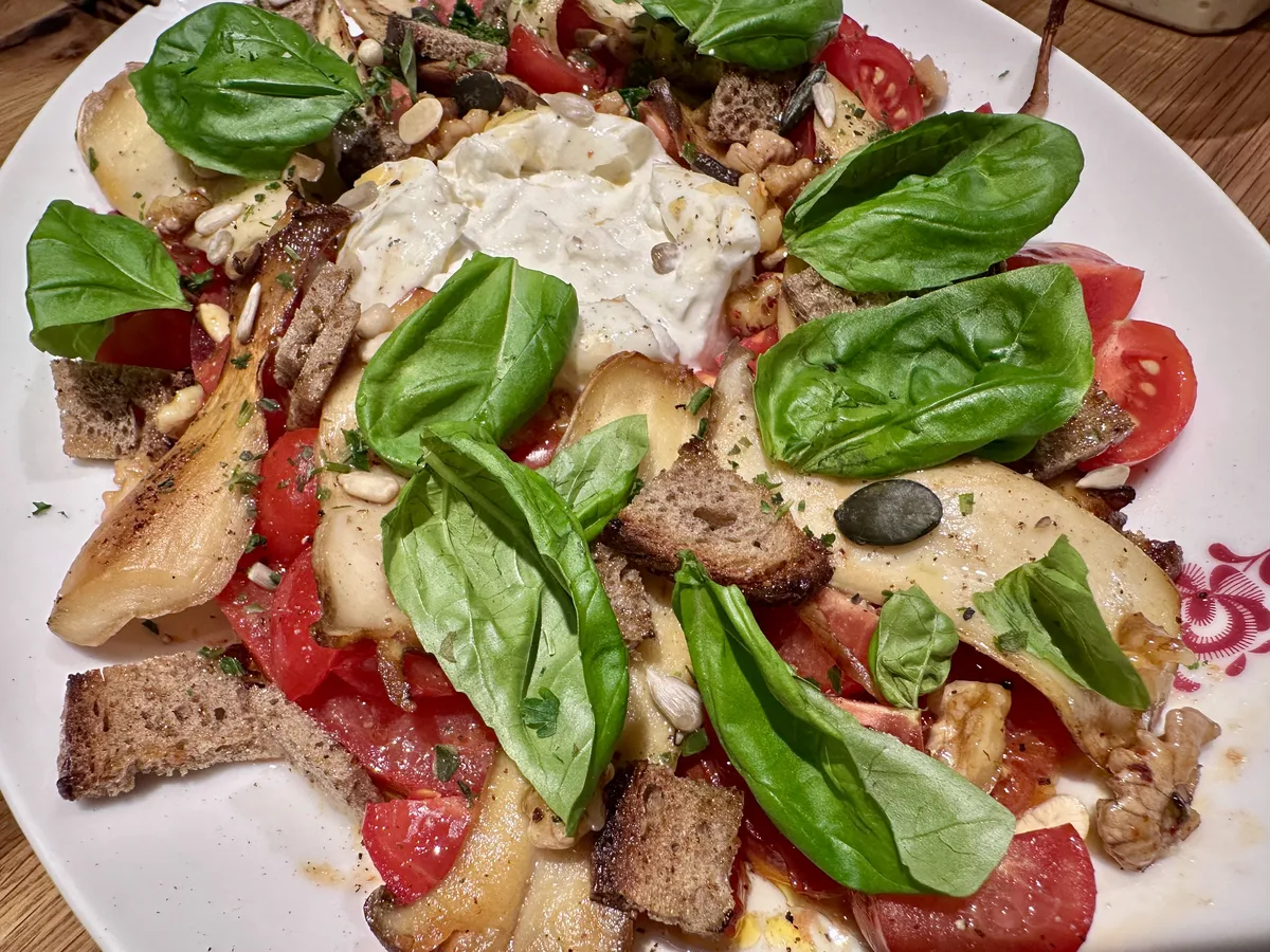 Ein Teller mit einem bunten Salat, Tomaten, geröstete Brotwürfel und frisches Basilikum enthält. In der Mitte befindet sich eine Portion cremiger Burrata, die das Gericht ansprechend ergänzt.
