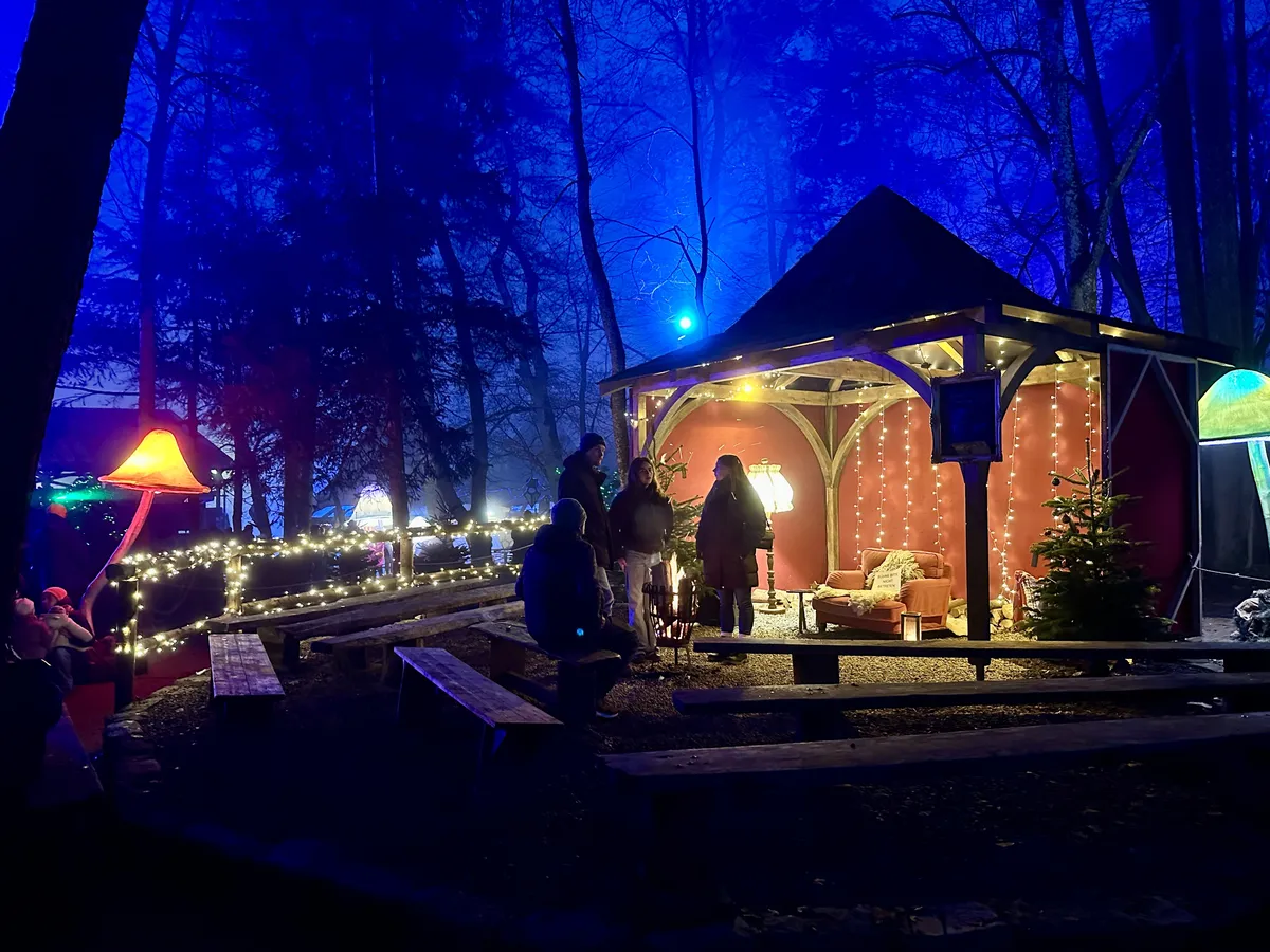 Eine nächtliche Szene im Freien zeigt einen hölzernen Pavillon, der mit Lichterketten, einem kleinen Weihnachtsbaum und einem Sessel dekoriert ist. Menschen versammeln sich im und um den Pavillon in einem blau beleuchteten Wald, mit beleuchteten pilzförmigen Dekorationen und Holzbänken im Vordergrund.