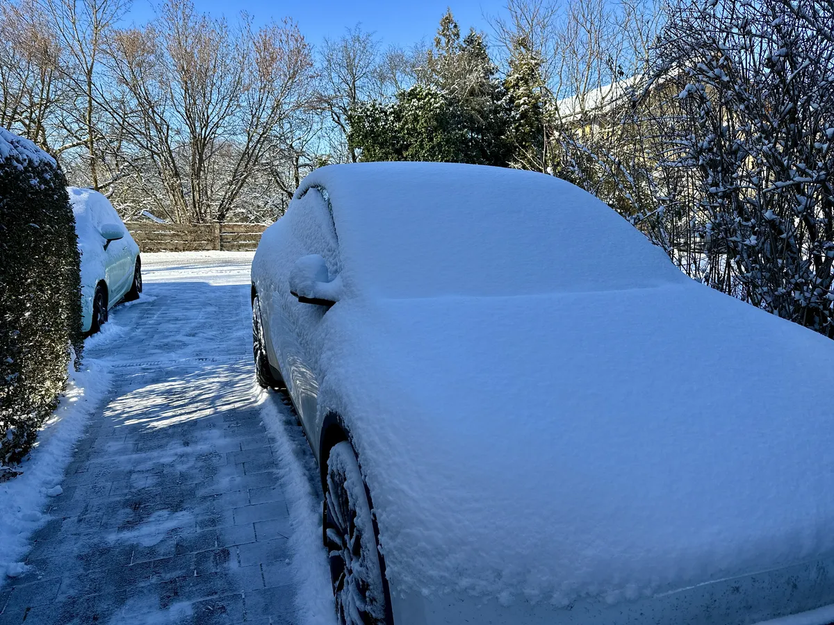 Ein mit Schnee bedecktes Auto steht auf einem schneebedeckten Parkplatz, umgeben von kahlen Bäumen und strahlend blauem Himmel. Der Schnee verdeckt die Konturen des Fahrzeugs vollständig.