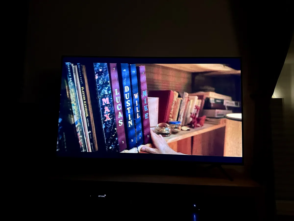 Ein Fernseher zeigt eine Nahaufnahme eines Bücherregals mit bunten Buchrücken, auf denen die Namen Lucas, Dustin, Max und Mike stehen. Eine Hand greift nach einem Buch im Regal.