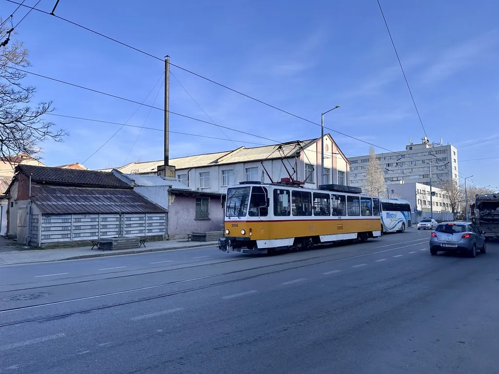 Eine gelbe Tatra-Straßenbahn fährt auf einem Straßenabschnitt, während im Hintergrund ein modernes Gebäude und mehrere andere Fahrzeuge sichtbar sind. Der Himmel ist klar und blau.
