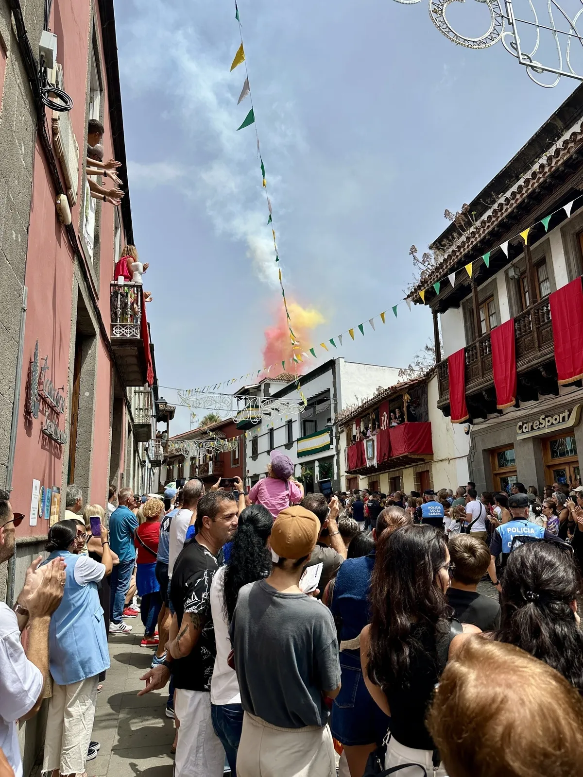 Eine Menschenmenge versammelt sich auf einer mit festlichen Girlanden geschmückten Straße und beobachtet ein Feuerwerk im Hintergrund.