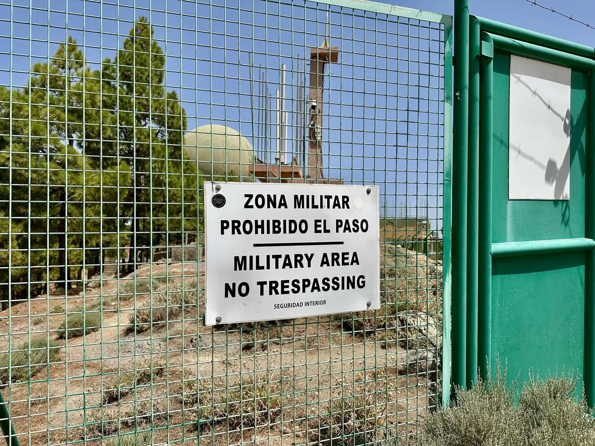 Ein Schild mit der Aufschrift Military Area - No Trespassing" hängt an einem Metallzaun, der ein Gelände mit militärischen Einrichtungen auf dem Pico de las Nieves umgibt.