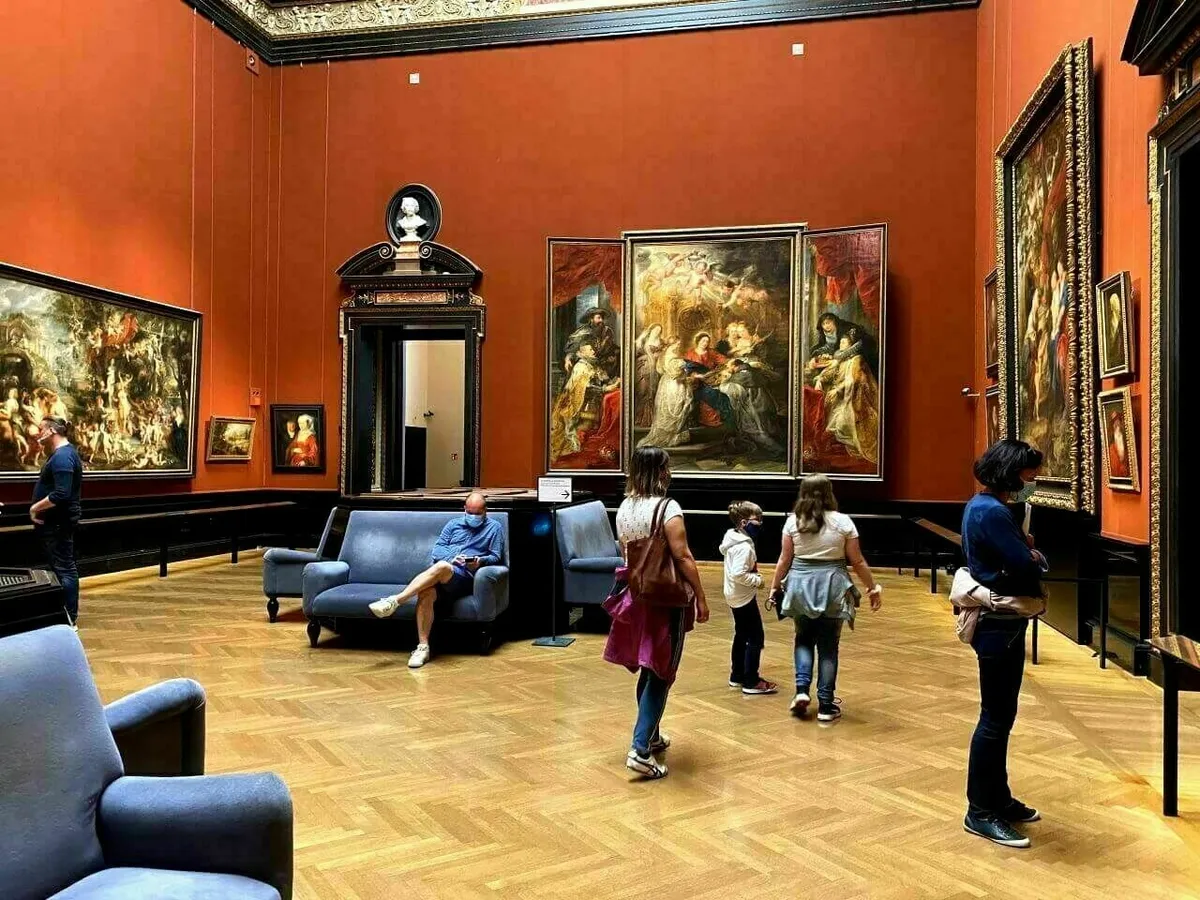 Das Innere des kunsthistorischen Museum Wien mit auffällig roten Wänden und einem Parkettboden. Mehrere Besucher betrachten große, gerahmte Ölgemälde, darunter ein zentrales Triptychon.