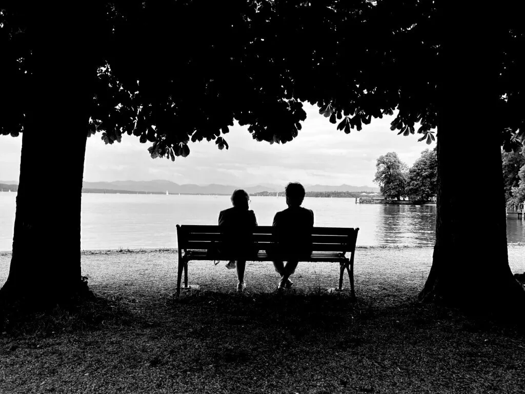 Zwei Damen sitzen auf einer Bank am Ufer des Starnberger Sees, umgeben von Bäumen, und blicken auf das Wasser.