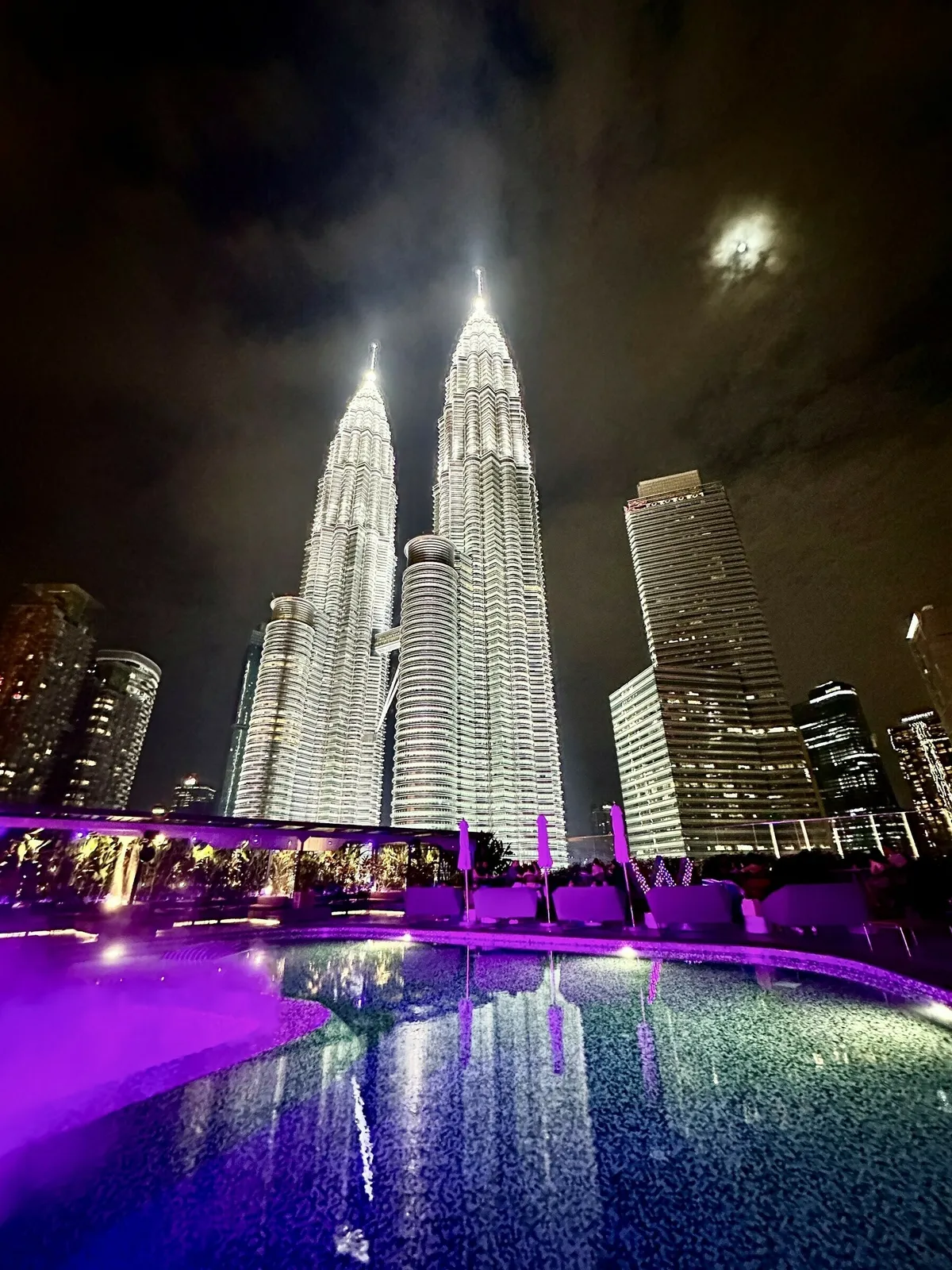 Die Petronas Twin Towers in Kuala Lumpur bei Nacht, hell erleuchtet gegen den dunklen Himmel und einen leuchtenden Vollmond. Im Vordergrund spiegeln sich die beleuchteten Türme in einem Swimmingpool mit starkem violettem Licht.