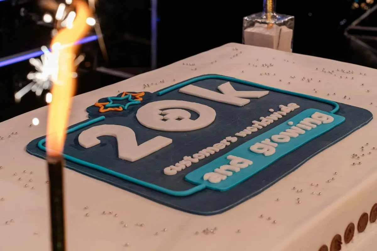 Nahaufnahme einer Torte, verziert mit der Aufschrift „20K customers worldwide and growing“ und einer brennenden Wunderkerze.
