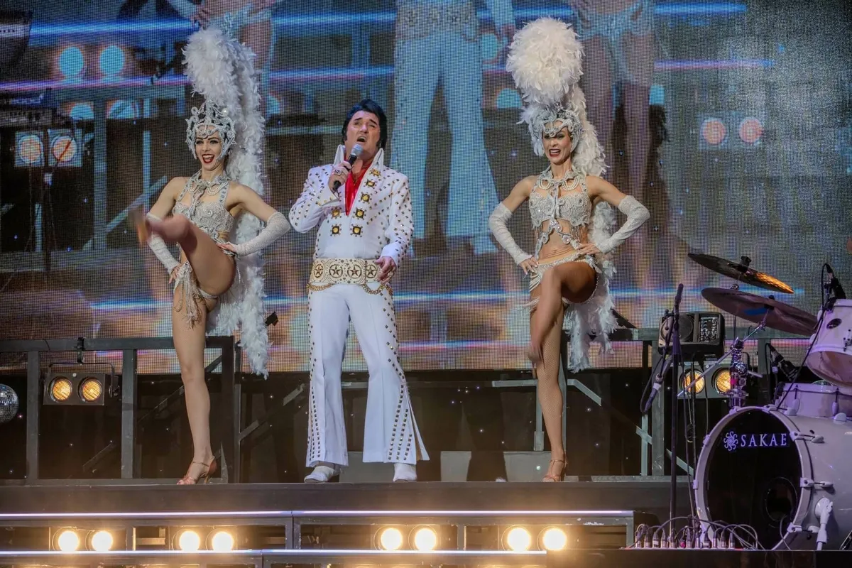 Ein Elvis-Imitator in weißem Outfit singt auf einer Bühne zwischen zwei Tänzerinnen mit Feder-Kopfschmuck.