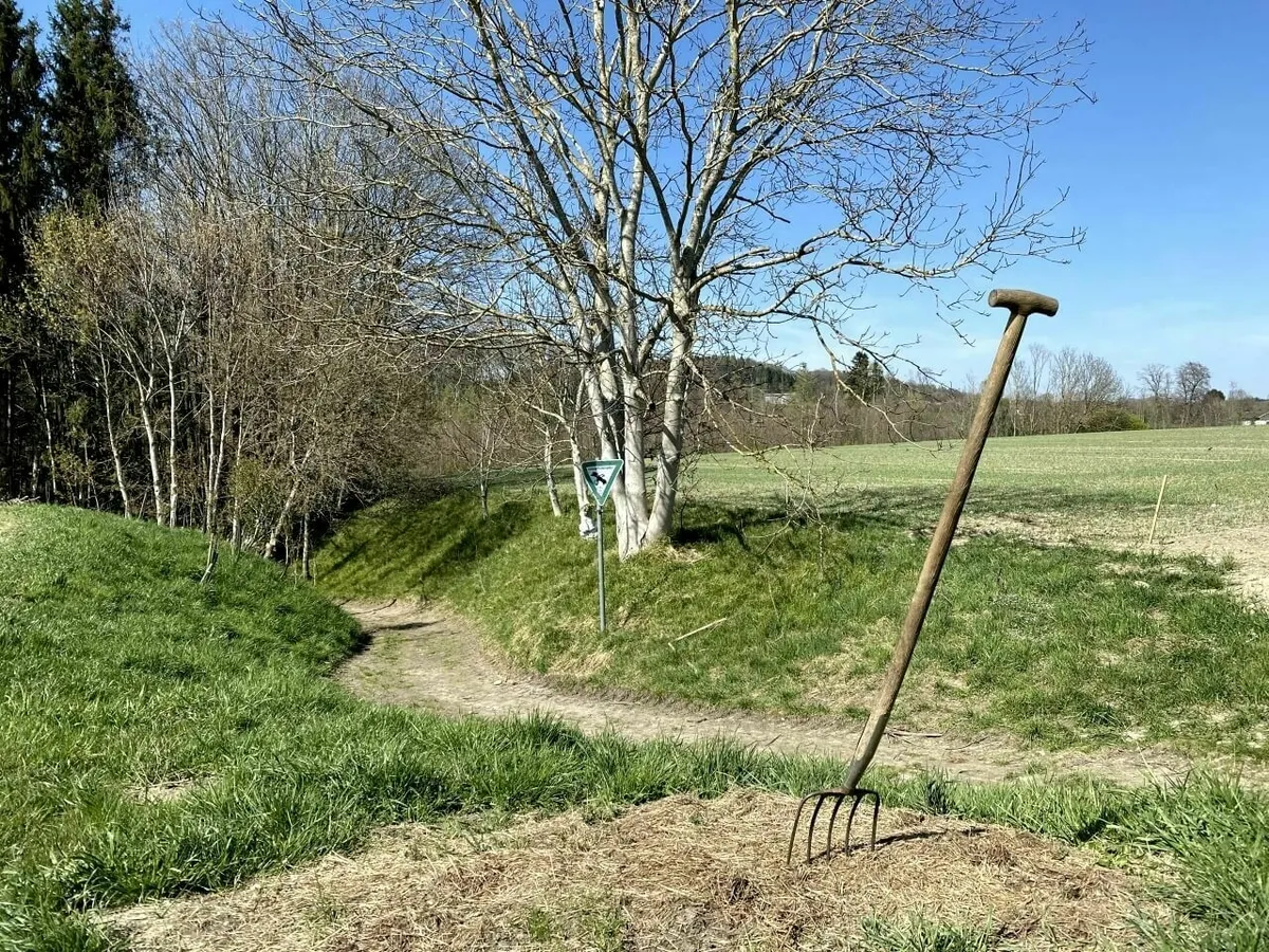 Eine hölzerne Mistgabel steckt in einem Haufen getrocknetem Material neben einem schmalen Wanderweg, umgeben von hohem Gras und einem kahlen Baum im Hintergrund.