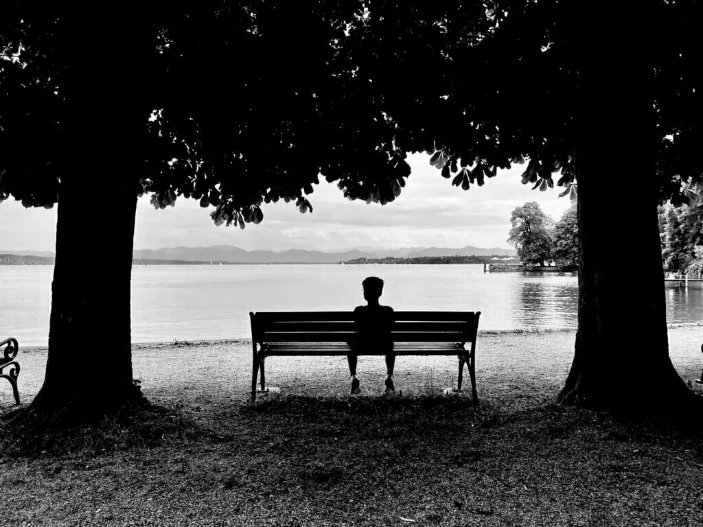 Schwarz-Weiß-Aufnahme, die eine einzelne Person in Silhouette auf einer Parkbank zwischen zwei großen Bäumen zeigt. Die Person blickt auf einen See mit Bergen im Hintergrund.