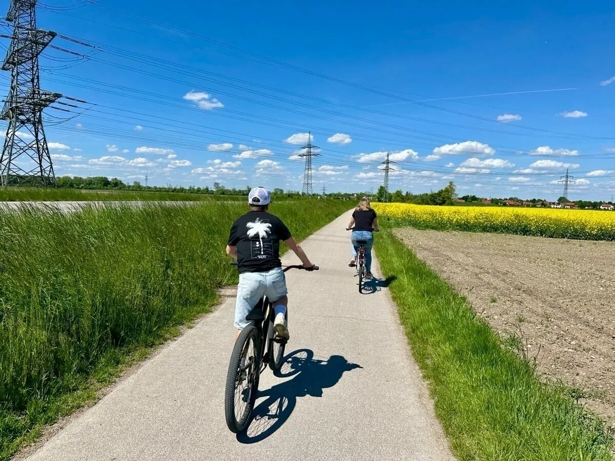 Zwei Personen fahren an einem sonnigen Tag auf einem asphaltierten Radweg durch die Natur. Die Person im Vordergrund trägt ein schwarzes T-Shirt, helle Jeans-Shorts und eine weiße Kappe und fährt auf einem Mountainbike. Die zweite Person ist etwas weiter entfernt, trägt dunkle Kleidung und fährt ebenfalls Rad. Der Weg führt durch hohes grünes Gras. Auf der rechten Seite ist ein leuchtend gelbes Rapsfeld zu sehen. Im Hintergrund dominieren mehrere hohe Strommasten die Landschaft unter einem strahlend blauen Himmel mit wenigen weißen Wolken.
