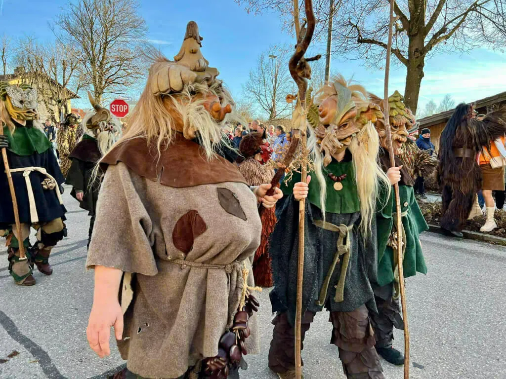 Zwei Personen in naturfarbenen, moos- und laubartigen Kostümen mit holzigen Gesichtsmasken und Stöcken nehmen am Umzug teil.