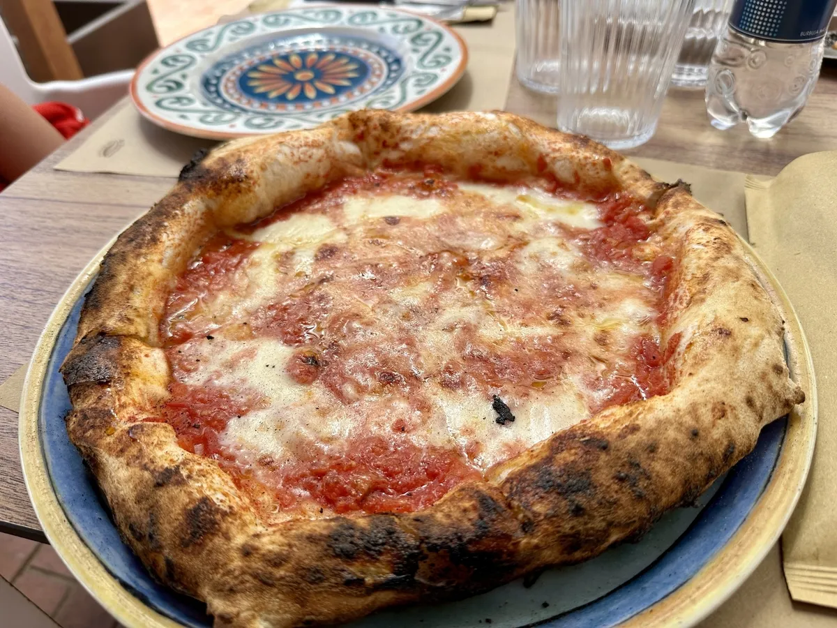 Eine Pizza mit Tomatensoße und geschmolzenem Käse liegt auf einem Teller auf einem Tisch, umgeben von Gläsern und einem anderen Teller mit Muster.