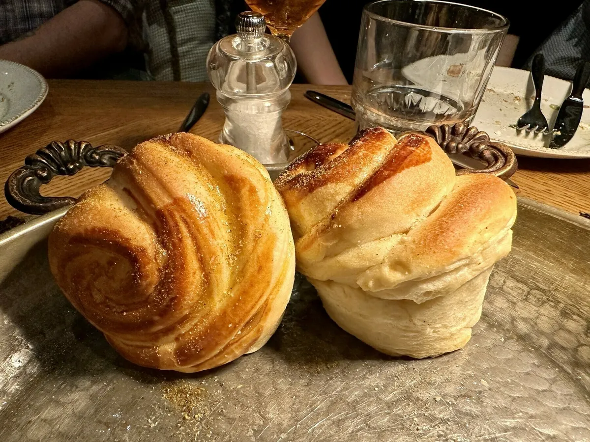 Zwei Zimtschnecken-artige, goldbraune Hefeteilchen auf einem metallischen Tablett, bestreut mit Gewürzen, auf einem Holztisch.