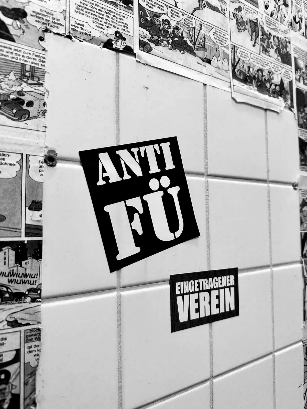 Schwarz-weißes Bild, das zwei Sticker auf weißen quadratischen Fliesen zeigt: Oben ein großer schwarzer Aufkleber mit weißer Schrift "ANTI FÜ" und darunter ein kleinerer, rechteckiger schwarzer Aufkleber mit weißer Schrift "EINGETRAGENER VEREIN". Die Fliesen sind teilweise mit Comic-Streifen beklebt.